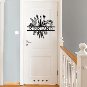 May include: Black metal wall art featuring a stylized design of paintbrushes, flowers, and leaves. The name "KIRKLAND" is displayed on a banner.