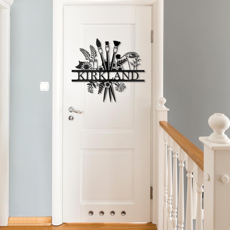 May include: Black metal wall art featuring a stylized design of paintbrushes, flowers, and leaves. The name "KIRKLAND" is displayed on a banner.
