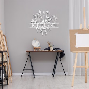 May include: White metal wall art featuring a stylized design of paintbrushes, paint tubes, and floral elements. The name "KIRKLAND" is displayed in a banner above the design.