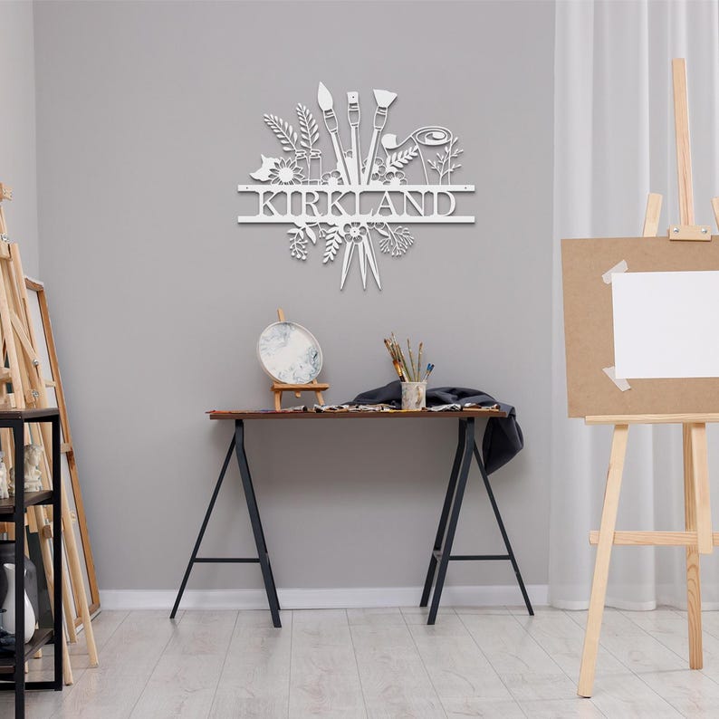 May include: White metal wall art featuring a stylized design of paintbrushes, paint tubes, and floral elements. The name "KIRKLAND" is displayed in a banner above the design.