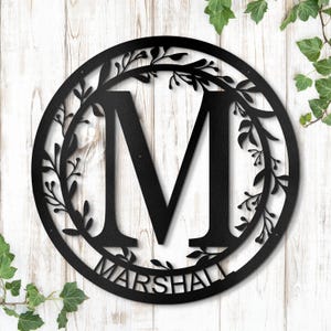 May include: Black metal monogram sign with a floral wreath design. The letter "M" is in the center of the wreath and the name "MARSHALL" is written below the letter.