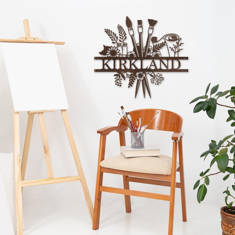 May include: A brown metal wall art piece with the name "KIRKLAND" in the center. The art features paintbrushes, flowers, and other artistic elements.