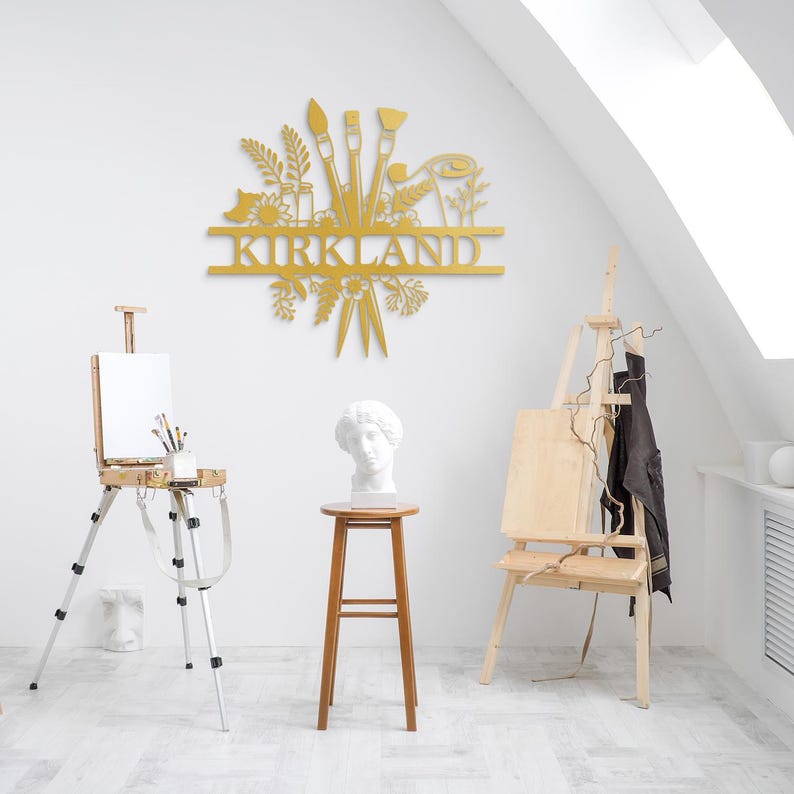 May include: Gold metal wall art featuring a stylized design of paintbrushes, flowers, and leaves. The name "KIRKLAND" is displayed in a banner below the design.