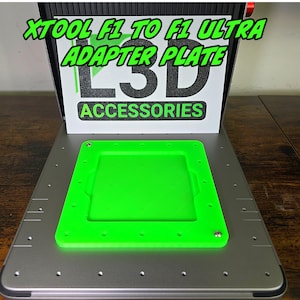 Puede incluir: Una placa adaptadora de plástico verde para una impresora 3D. La placa tiene un recorte rectangular en el centro y está etiquetada como "XTOOL F1 TO F1 ULTRA ADAPTER PLATE" y "L3D ACCESSORIES".