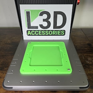 Op de afbeelding: Een groene plastic bouwplaat met een rechthoekige uitsparing en meerdere gaten. De bouwplaat ligt op een zilveren metalen oppervlak. De tekst "L3D ACCESSORIES" is zichtbaar op een wit bord achter de bouwplaat.