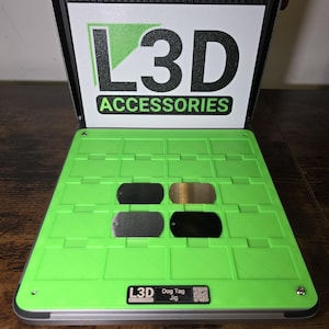 May include: A green dog tag jig with four metal dog tags. The jig has the text "L3D ACCESSORIES" and "Dog Tag Jig". The dog tags are black, silver, and gold.
