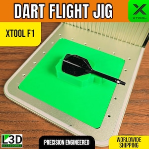 Puede incluir: Una plantilla de vuelo de dardos con el texto "DART FLIGHT JIG" y "XTOOL F1". La plantilla tiene un marco plateado, un centro verde brillante y una pluma de dardo negra. También se ve el texto "PRECISION ENGINEERED".