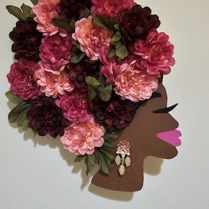 Könnte beinhalten: Wanddekoration mit dem Profil einer Frau, geschmückt mit einem Blumenarrangement in Rosa- und Burgundertönen. Grüne Blätter ergänzen die Blumen. Die Lippen der Frau sind rosa bemalt, und sie trägt goldene Ohrringe.