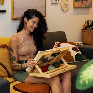 May include: A woman is holding a wooden embroidery frame with red and green yarn. She is sitting on a gray couch with yellow and brown throw pillows. The wall behind her is decorated with framed artwork and a small sign that says "CIAO".