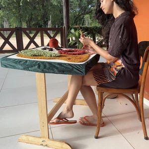 Pode incluir: Uma pessoa está trabalhando em um projeto de crochê de tapete. O tapete está em uma mesa pequena com uma borda de tecido verde. O tapete tem seções de fio verde, preto, vermelho e amarelo. Uma bola de fio laranja está na mesa.