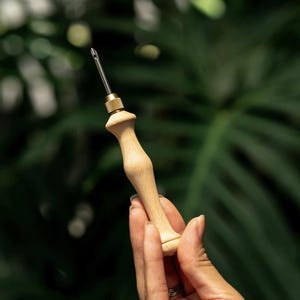 Pode incluir: Uma ferramenta de agulha de perfuração de madeira com uma conexão de latão e uma agulha prateada. A alça é de madeira clara e a agulha se estende da parte superior. A ferramenta é projetada para projetos de crochê e bordado.