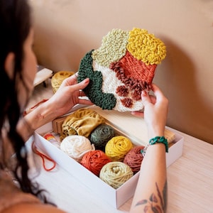 Pode incluir: Uma colorida peça de arte de agulha mágica com secções verdes, amarelas, laranja e brancas é segurada acima de uma caixa branca cheia de novelos de lã. Os novelos de lã são de várias cores, incluindo branco, amarelo, laranja e verde.
