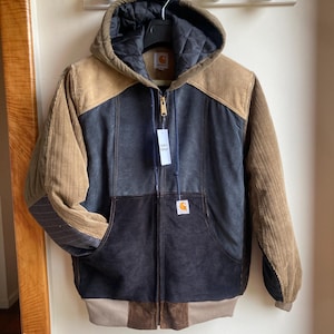 May include: A hooded Carhartt jacket with a zip-up front. The jacket features a color-block design with corduroy and quilted panels in shades of brown, tan, and navy blue. A Carhartt label is visible on the chest. The jacket is hanging on a wall.