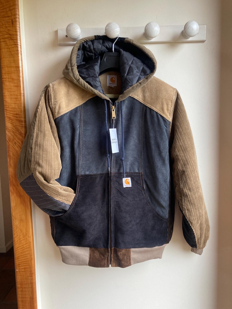 May include: A hooded Carhartt jacket with a zip-up front. The jacket features a color-block design with corduroy and quilted panels in shades of brown, tan, and navy blue. A Carhartt label is visible on the chest. The jacket is hanging on a wall.