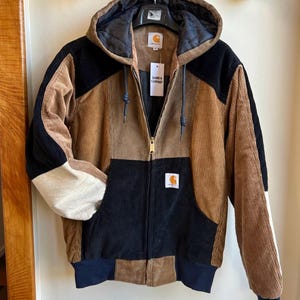 Op de afbeelding: Een corduroy jack met capuchon in een color-block ontwerp, voorzien van een volledige ritssluiting, geribbelde boorden en een capuchon met trekkoord. Het kledingstuk heeft een patchworkpatroon in beige, zwart en crème met een Carhartt-label.