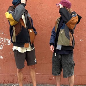 May include: Two people wearing patchwork jackets with multiple colors, shorts, and hats. One person wears brown sandals, and the other wears green sneakers. The background is a textured, reddish-orange wall.