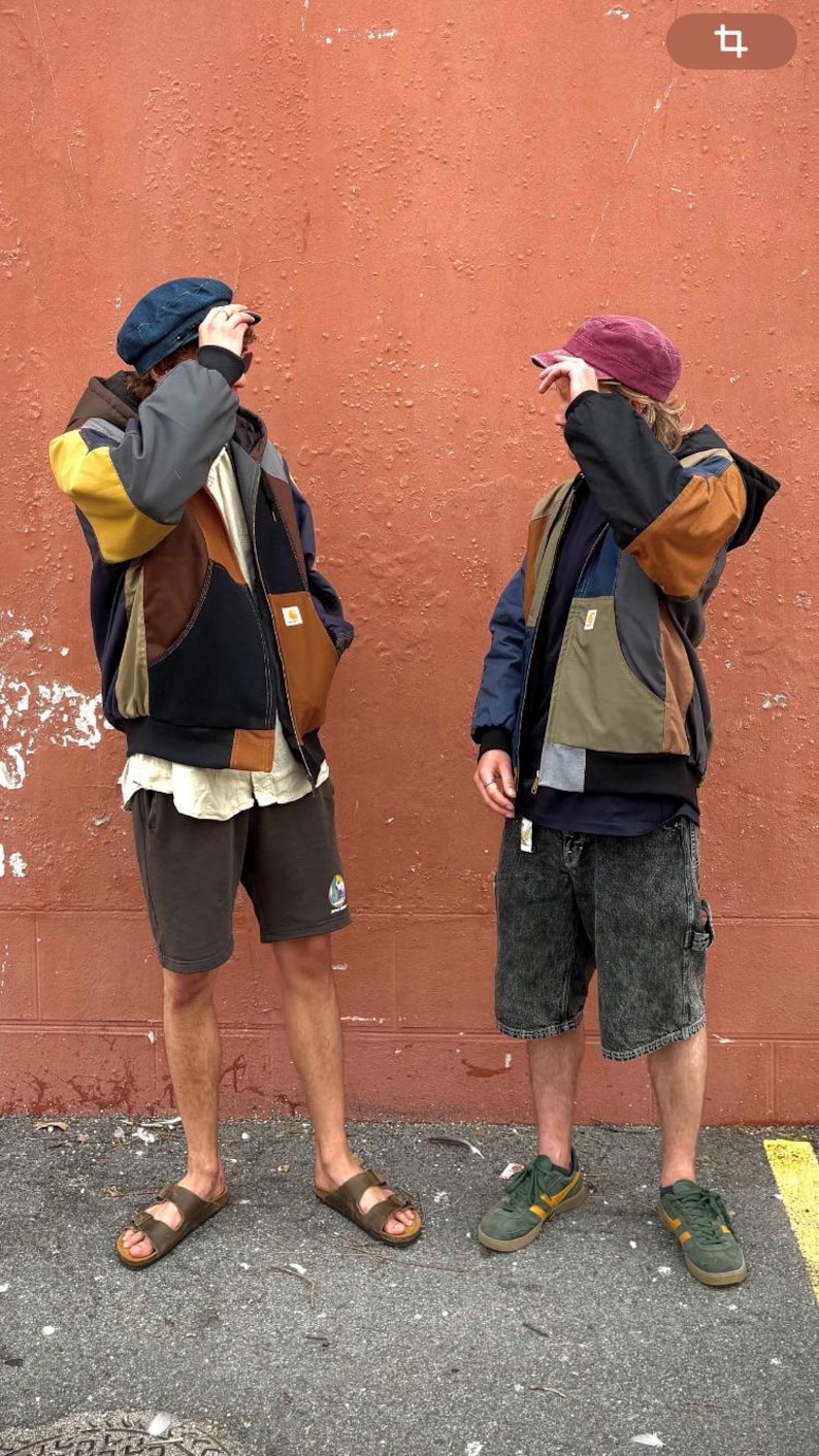 May include: Two people wearing patchwork jackets with multiple colors, shorts, and hats. One person wears brown sandals, and the other wears green sneakers. The background is a textured, reddish-orange wall.