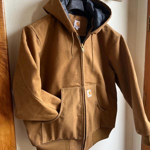 Pode incluir: Casaco com capuz castanho com forro interior preto e zíper dourado. O casaco tem dois bolsos frontais e um patch com o logotipo Carhartt. Pendurado numa parede branca.