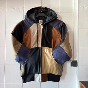 May include: A patchwork hooded jacket with a zip-up front. The jacket features panels of black, brown, tan, and blue fabric. It has a black ribbed hem and cuffs, and a black hood. A small Carhartt logo is visible on the front.