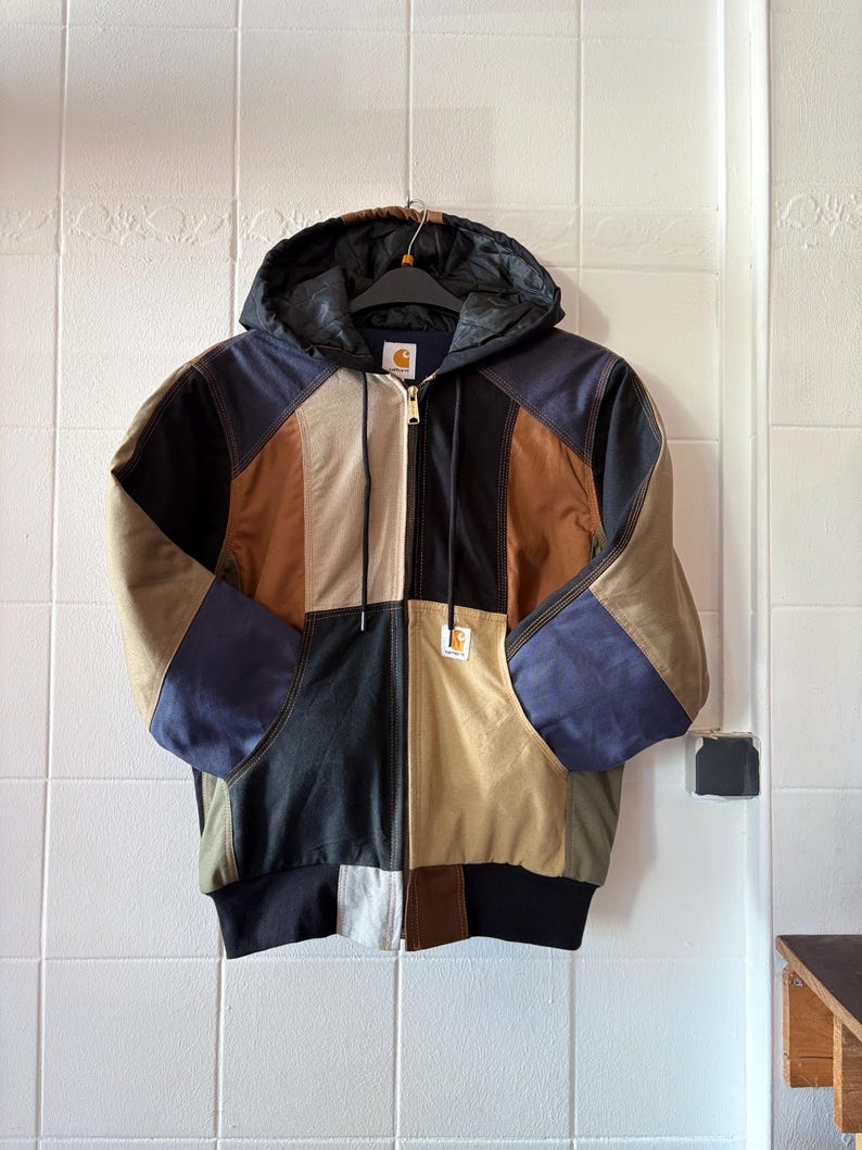 May include: A patchwork hooded jacket with a zip-up front. The jacket features panels of black, brown, tan, and blue fabric. It has a black ribbed hem and cuffs, and a black hood. A small Carhartt logo is visible on the front.