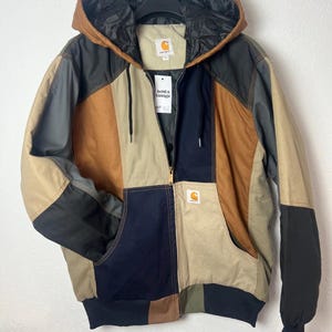 May include: A colour-blocked hooded jacket featuring panels of tan, brown, navy blue, and charcoal grey. The jacket has a front zip closure, a drawstring hood, ribbed cuffs, and a Carhartt brand patch on the chest pocket and inner neck label.