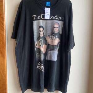 May include: Black t-shirt featuring a graphic of two men with sunglasses and the text "The Outsiders." The shirt also has the text "nWo" on the bottom. The shirt is hanging on a black hanger.