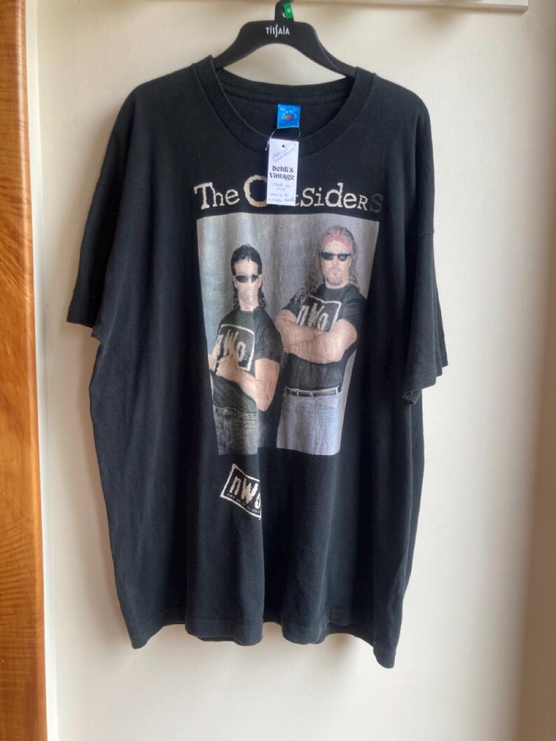 May include: Black t-shirt featuring a graphic of two men with sunglasses and the text "The Outsiders." The shirt also has the text "nWo" on the bottom. The shirt is hanging on a black hanger.