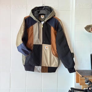 May include: A multi-coloured patchwork hooded jacket with a zip-up front. The jacket features black, brown, tan, and navy blue panels. The Carhartt logo is visible on the front pocket. The jacket is hanging on a white tiled wall.