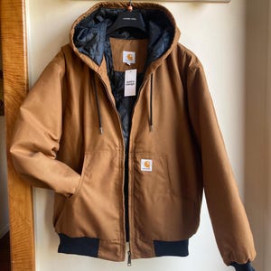 Könnte beinhalten: Braune Kapuzenjacke mit schwarzem Innenfutter und schwarzen gerippten Bündchen. Die Jacke hat einen Reißverschluss und zwei Vordertaschen. Ein weißes Etikett mit schwarzem Text ist angebracht. Das Carhartt-Logo ist sichtbar.