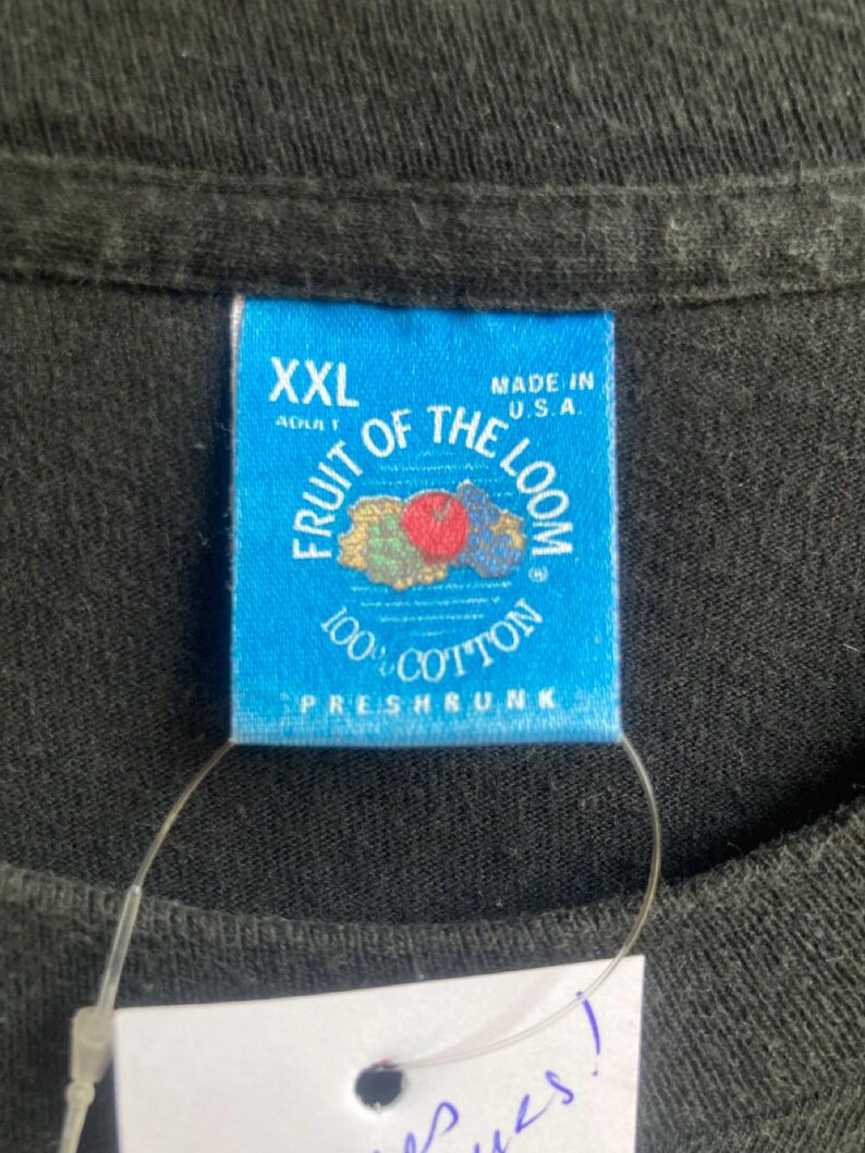 May include: Close-up of a black t-shirt with a blue tag. The tag reads "XXL ADULT" and "FRUIT OF THE LOOM 100% COTTON PRESHRUNK MADE IN U.S.A." The tag has a fruit graphic.