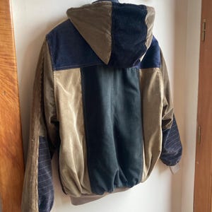 May include: A corduroy jacket with a patchwork design in shades of brown, navy, and black. The jacket features a hood and ribbed cuffs. The jacket is hanging on a white wall.