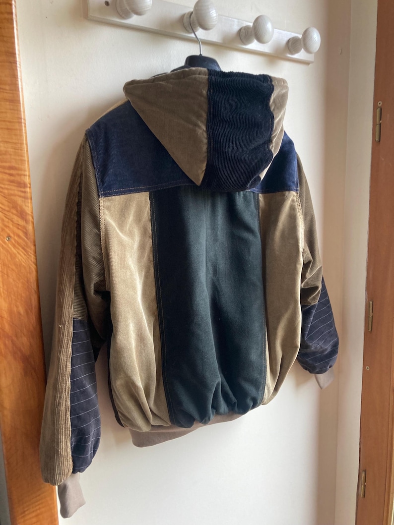 May include: A corduroy jacket with a patchwork design in shades of brown, navy, and black. The jacket features a hood and ribbed cuffs. The jacket is hanging on a white wall.