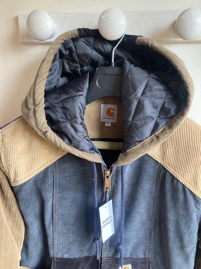 May include: A Carhartt corduroy jacket in shades of brown, tan, and blue, with a quilted black hood lining. The jacket has a full zipper, a tag that reads "beldi's vintage", and a Carhartt label.