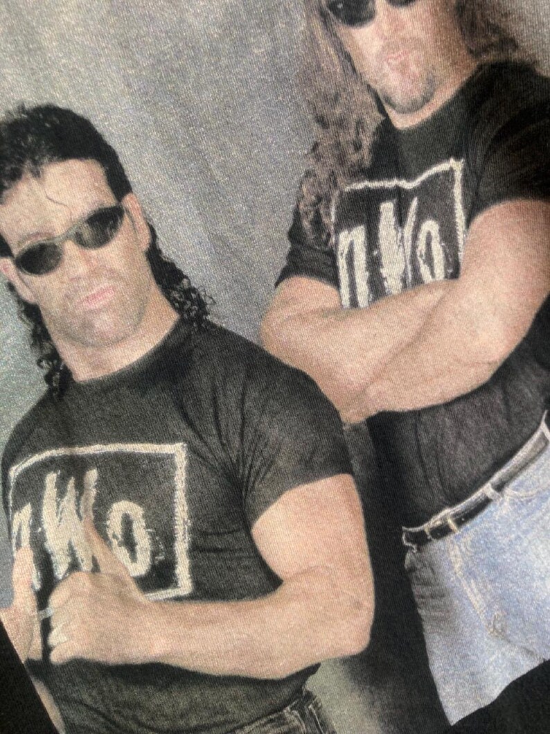 May include: Two men wearing black t-shirts with the letters "nWo" in a white box, and sunglasses. The men are standing close together, with one giving a thumbs up. The background is a light gray color.