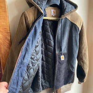May include: A multi-toned corduroy jacket with a hood, featuring brown, blue, and black panels. The jacket has a quilted black lining and a Carhartt logo patch. It is hanging on a hanger.