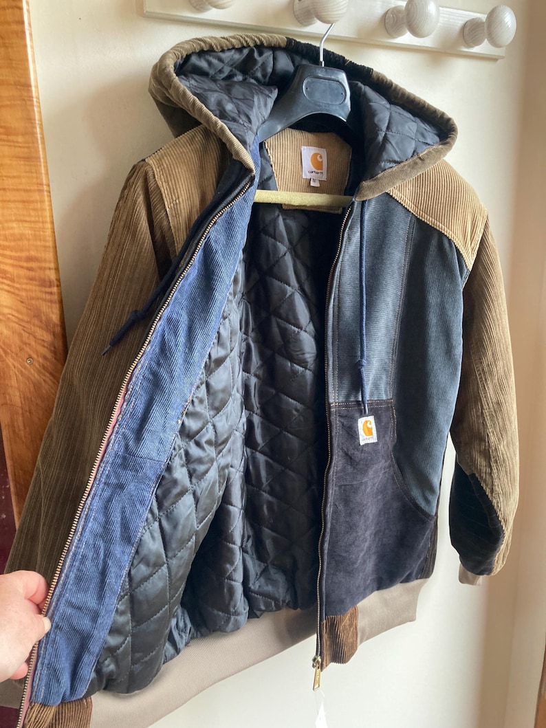 May include: A multi-toned corduroy jacket with a hood, featuring brown, blue, and black panels. The jacket has a quilted black lining and a Carhartt logo patch. It is hanging on a hanger.