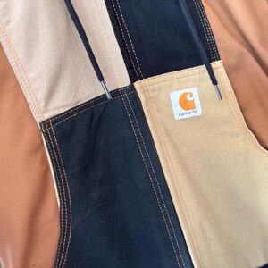 May include: Close-up of a multi-colored jacket with a patchwork design. The jacket features panels of tan, black, and brown fabric, with visible stitching. A small square label with an orange logo is sewn on the jacket.