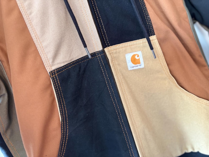 May include: Close-up of a multi-colored jacket with a patchwork design. The jacket features panels of tan, black, and brown fabric, with visible stitching. A small square label with an orange logo is sewn on the jacket.