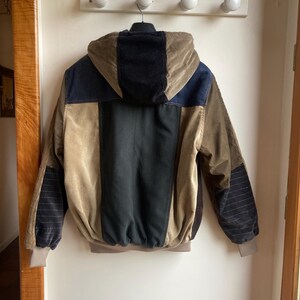 May include: A multi-colored corduroy jacket with a hood. The jacket features panels of black, navy blue, and tan corduroy. The sleeves have a striped pattern. The jacket is hanging on a white wall.