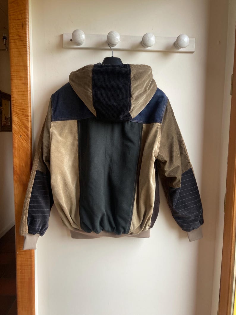 May include: A multi-colored corduroy jacket with a hood. The jacket features panels of black, navy blue, and tan corduroy. The sleeves have a striped pattern. The jacket is hanging on a white wall.
