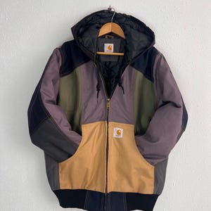 Op de afbeelding: Een color-block jack met capuchon voorzien van panelen in beige, olijfgroen, donkergrijs en zwart. Het jack heeft een ritssluiting aan de voorzijde, een gewatteerde zwarte voering en een Carhartt-merklabel op de borstzak en in de nek.