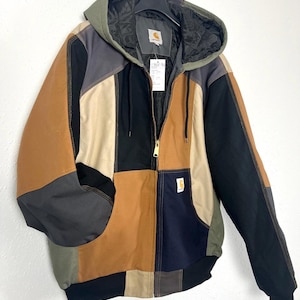 Könnte beinhalten: Eine farblich abgesetzte Kapuzenjacke mit Einsätzen aus beigem, schwarzem, dunkelgrauem und olivgrünem Stoff. Die Jacke verfügt über einen Frontreißverschluss, ein schwarzes Steppfutter, eine Kapuze mit Kordelzug sowie ein Carhartt-Logo auf Brust und Kragen.