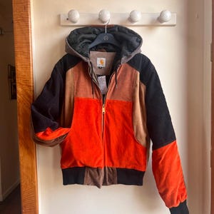 Op de afbeelding: Een color-block jack met capuchon, bestaande uit panelen van oranje, bruine en zwarte corduroy stof. Het jack heeft een ritssluiting aan de voorzijde, een donkergrijze capuchon met trekkoorden, een geribbelde zoom en een Carhartt-label in de nek.