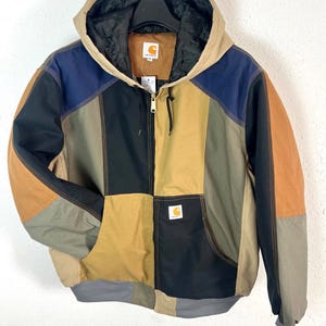 Könnte beinhalten: Eine farblich abgesetzte Kapuzenjacke mit Patchwork-Design in den Farben Beige, Schwarz, Marineblau und Olivgrün. Die Jacke verfügt über einen Frontreißverschluss, ein schwarzes Steppfutter und ein Carhartt-Markenlogo auf der Brusttasche.