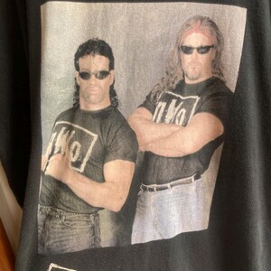 May include: Black t-shirt featuring two men wearing sunglasses, with the text "The Outsiders" above a photo. The men are wearing black t-shirts with the "nWo" logo. The words "new world order" are below the logo.