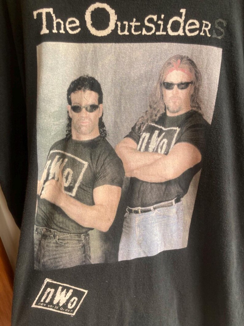 May include: Black t-shirt featuring two men wearing sunglasses, with the text "The Outsiders" above a photo. The men are wearing black t-shirts with the "nWo" logo. The words "new world order" are below the logo.