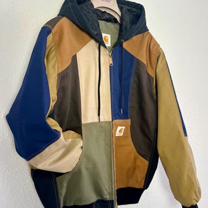 May include: A colour-blocked hooded jacket featuring panels of navy blue, tan, olive green, and dark brown. The jacket has a front zip closure, a black hood with drawstrings, black ribbed cuffs and hem, and a Carhartt brand patch on the chest.