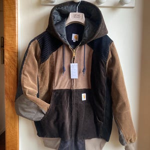 Peut inclure: Veste en velours côtelé multicolore avec capuche. Elle présente un assemblage de panneaux marron, noir et beige, une fermeture éclair et un petit écusson logo. La veste est suspendue à un porte-manteau blanc avec des boutons en bois. L'étiquette indique "MEME ROAD".