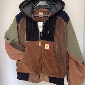 Op de afbeelding: Een color-blocked corduroy jas met capuchon, voorzien van een volledige ritssluiting, geribbelde boorden en zoom, en een zwarte doorgestikte voering. De jas heeft een patchworkontwerp in tinten bruin, olijfgroen en zwart met Carhartt-labels.