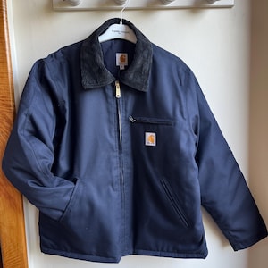 Op de afbeelding: Marineblauwe werkjas met een corduroy kraag en een gouden rits. De jas heeft twee voorzakken en een kleine logopatch op de borst. De jas hangt aan een witte hanger.